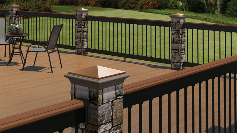 A composite deck with Deckorators Aluminum Railing and Gray Cobblestone Cast Stone Post Covers and Faux Stone Column Wraps is shown in front of a green lawn and rosebushes.