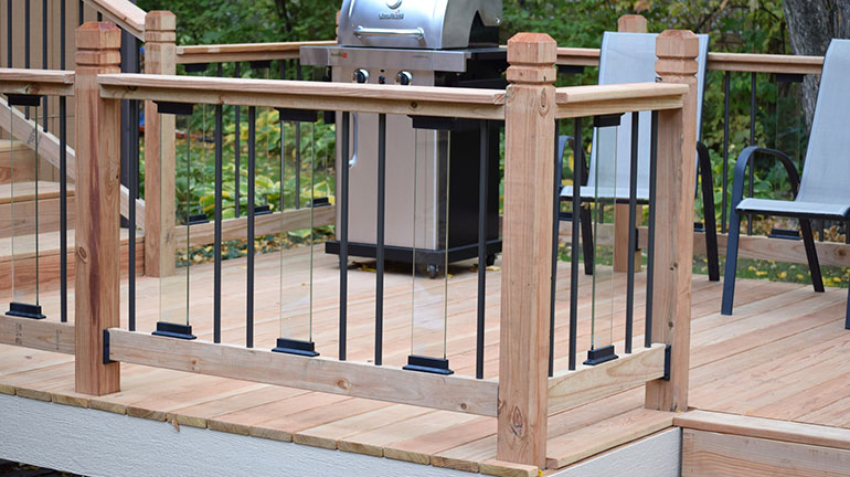 A cedar deck railing with glass and aluminum balusters and a grill next to the house in the background