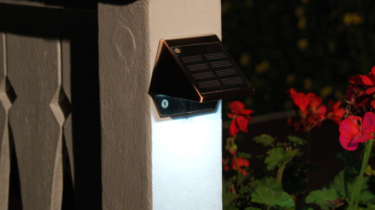 A copper-colored solar rail light from Classy Caps illuminates a white deck post and rail with red flowers in the background
