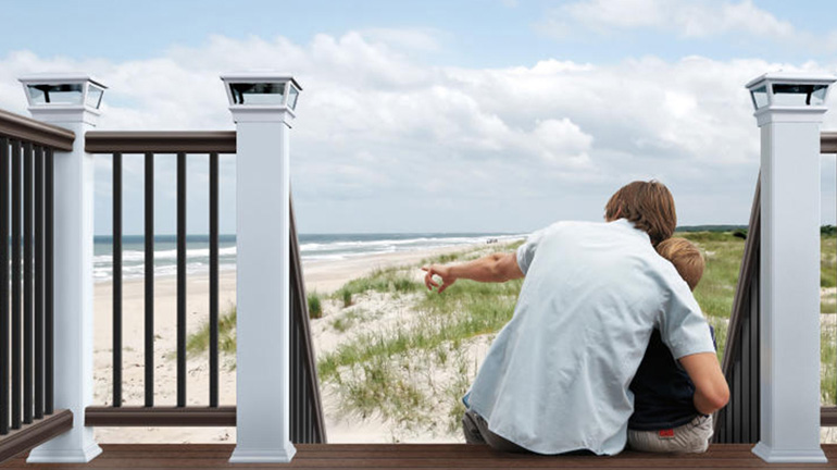 A man and boy sit on composite deck stairs leading down to a beach, the deck railing features white Deckorators post sleeves with Solar VersaCaps
